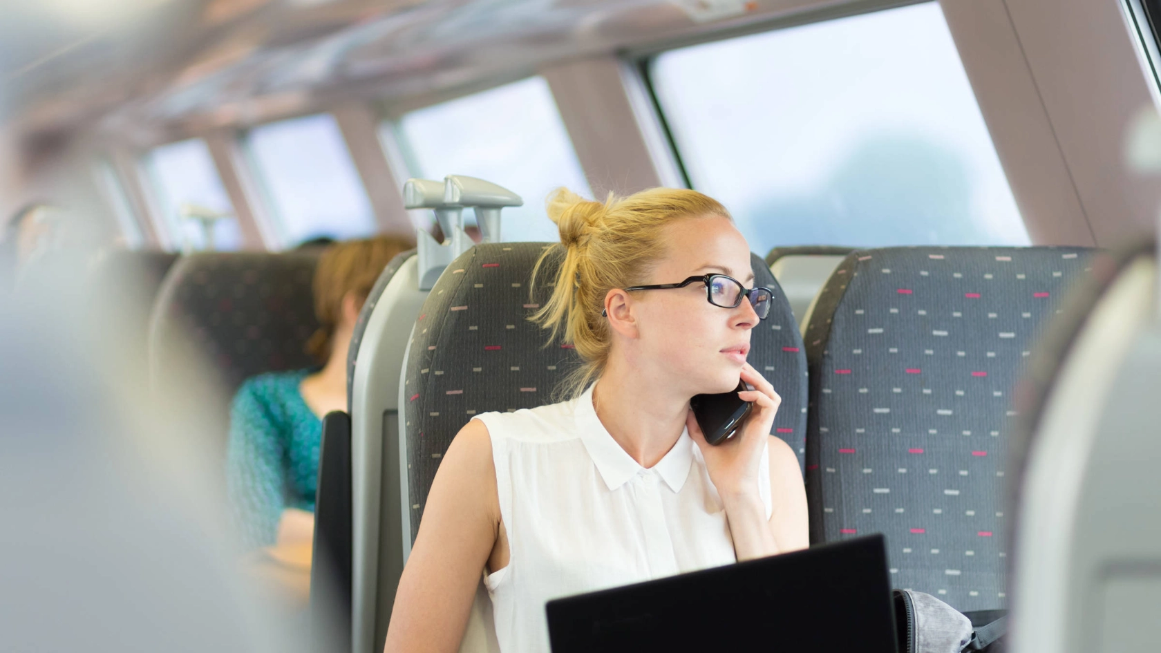 Eine junge Frau sitzt mit einem Notebook auf den Knien im Zug und telefoniert.