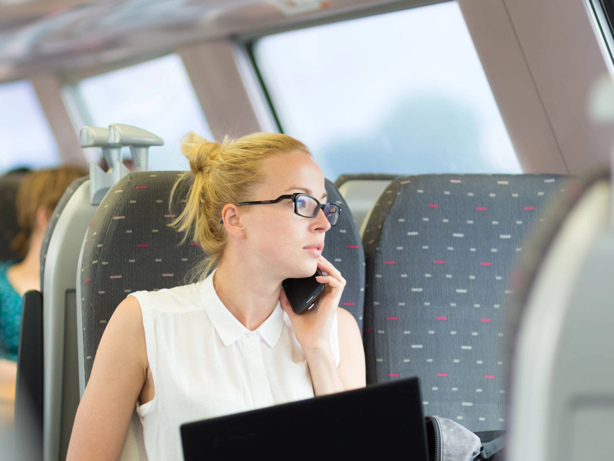 Eine junge Frau sitzt mit einem Notebook auf den Knien im Zug und telefoniert.